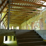 Zdjęcie przedstawia widok ze schodów prowadzących na drugie piętro budynku. Przy suficie widoczne są drewniane bale. Schody wykonane są z betonu. Ściany zbudowane są z cegły o kolorze pomarańczowym. W ścianach zlokalizowane są otwory na drzwi do małych pomieszczeń (w planach miały być one klasami). Pomieszczenie oświetla naturalne słońce padające z otworów znajdujących się z lewej strony zdjęcia, które przeznaczone są na okna.