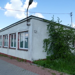 Zdjęcie przedstawia budynek starej szkoły w Chotyczach. Ma ona jasną wpadającą w biel elewację. Zdjęcie przedstawia budynek widoczny z wjazdu. Na budynku zamontowana jest latarnia. Wjazd ułożony jest z kostki brukowej szaro-czerwonej.