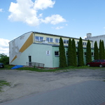 Zdjęcie przedstawia nowy, dobudowany budynek szkoły. Ma elewację koloru zielono-żółtego. Przed budynkiem od wewnętrznej strony rosną tuje.